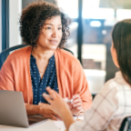 A middle aged mixed-race woman in a peach blazer with curly dark hair interviewing with a white woman wearing a light plaid shirt, who has straight dark hair